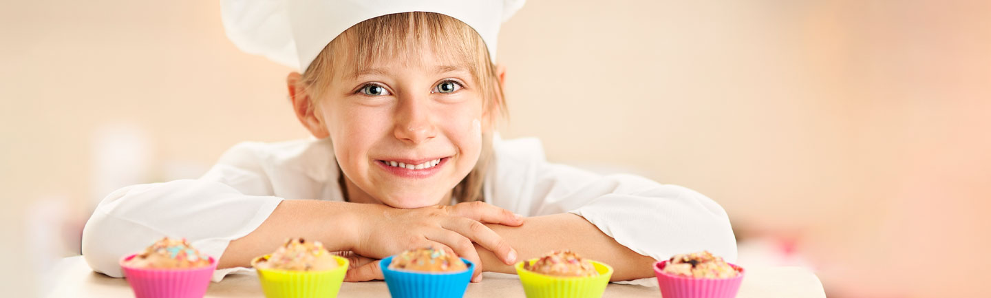 Young child with chef hat on proudly standing behind some cupcakes