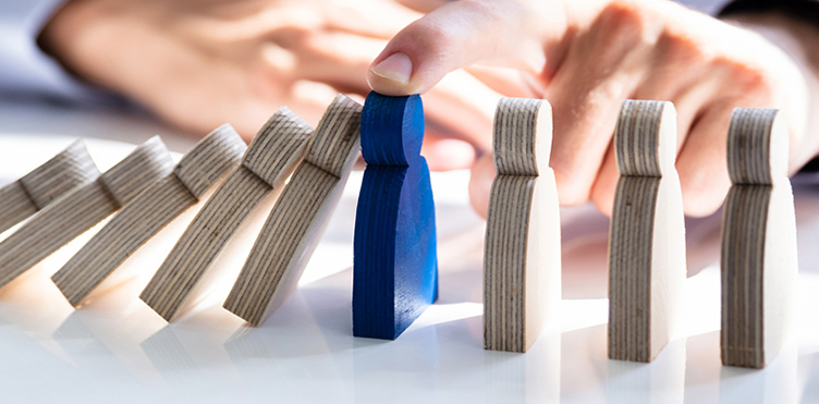 This photo is a close-up of a hand stopping the domino effect of wooden cutouts of people from falling on a desk