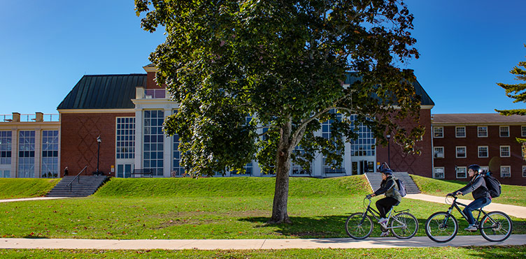 Two people bike along a path in front of trees and UNB campus buildings