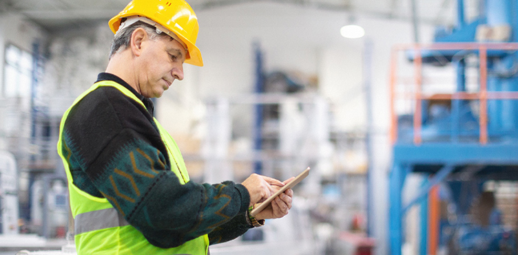 Warehouse technician wearing a hard hat checks inventory data on a tablet.