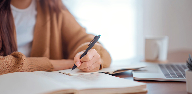 Photograph of a student writing with her laptop next to them on the desk